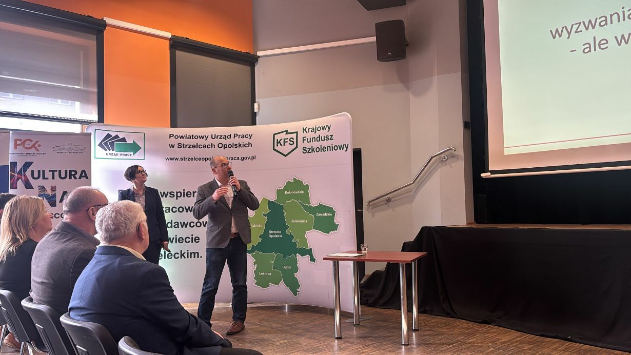 Na zdjęciu widać dwoje prelegentów – mężczyznę w szarej marynarce gestykulującego podczas mówienia oraz stojącą obok kobietę w ciemnym żakiecie. W tle wyświetlany jest fragment prezentacji dotyczący wyzwań rekrutacyjnych. Widoczna jest również ścianka promocyjna z hasłem "Razem wspieramy...". Publiczność obserwuje wystąpienie z krzeseł ustawionych w rzędach.