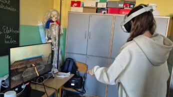 Uczennica w jasnej bluzie stoi tyłem do obiektywu, używając gogli VR w kąciku technologicznym klasy. Na monitorze przed nią widać wirtualny warsztat z kowadłem i narzędziami. W tle widać szkolne szafki, na których poukładane są liczne pudełka po butach.