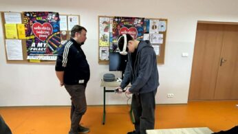 : Mężczyzna w sportowej bluzie rozmawia z uczniem, który ma na głowie gogle VR i patrzy w dół na kontrolery w swoich dłoniach. W tle na tablicy korkowej widoczne są plakaty Fundacji Wielka Orkiestra Świątecznej Pomocy (WOŚP).