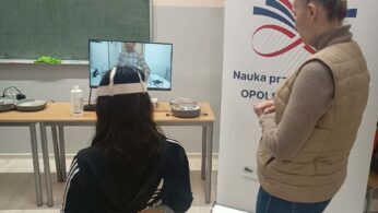 Uczennica siedzi na krześle tyłem do obiektywu, mając na głowie gogle VR. Przed nią na monitorze widać postać z symulacji. Obok stoi kobieta w beżowej kamizelce, która nadzoruje przebieg ćwiczenia. Po prawej stronie widoczny jest duży roll-up promujący projekt 