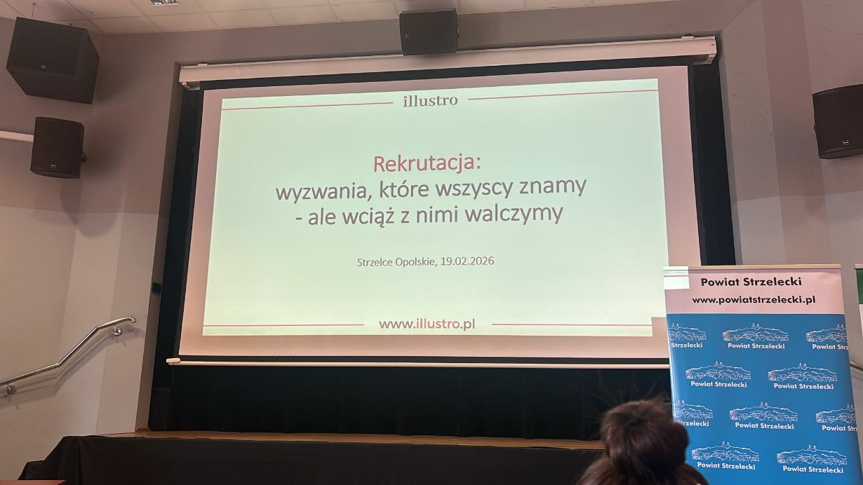 Bliskie ujęcie ekranu projekcyjnego z tytułowym slajdem prezentacji firmy "illustro". Napis na slajdzie brzmi: "Rekrutacja: wyzwania, które wszyscy znamy - ale wciąż z nimi walczymy". Pod spodem widnieje data 19.02.2026 oraz nazwa miejscowości Strzelce Opolskie. Po prawej stronie kadru widoczny jest fragment niebieskiego baneru z herbem powiatu.