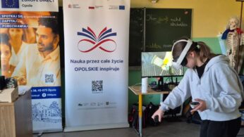 Uczennica pochyla się, korzystając z zestawu VR, co sugeruje interakcję z obiektem znajdującym się nisko w wirtualnej przestrzeni. Stoi przed banerem z napisem „Nauka przez całe życie OPOLSKIE inspiruje”. Na małym monitorze obok widoczny jest jasny, laboratoryjny widok z aplikacji.