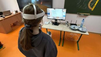 Widok z góry na uczennicę w goglach VR stojącą przed małym monitorem, na którym wyświetlana jest prezentacja z tekstem. Obok na ławce znajduje się router, butelka wody oraz otwarte etui na gogle. Na zielonej tablicy w tle widnieją napisy kredą i przyczepiony duży kątomierz.