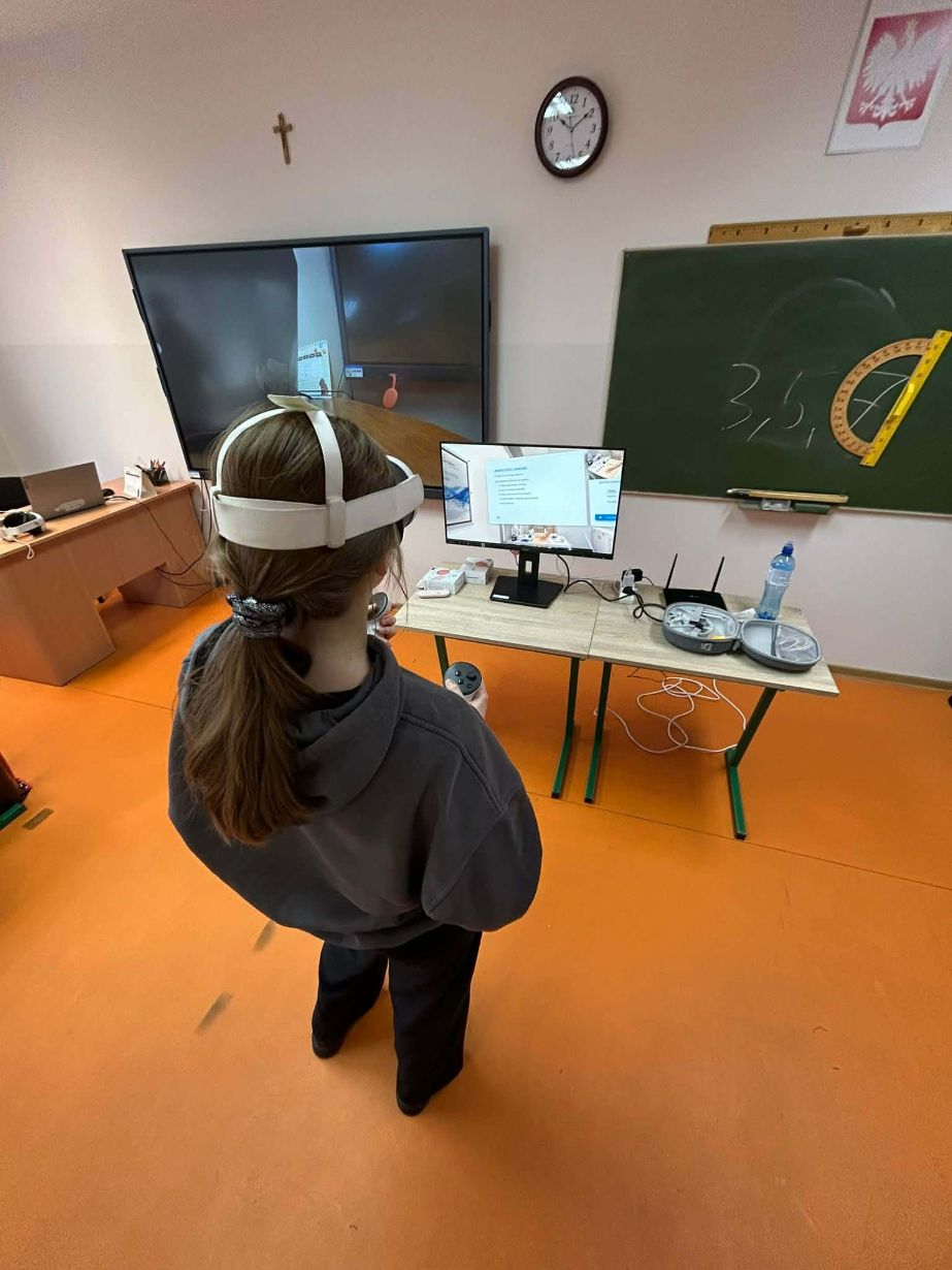 Widok z góry na uczennicę w goglach VR stojącą przed małym monitorem, na którym wyświetlana jest prezentacja z tekstem. Obok na ławce znajduje się router, butelka wody oraz otwarte etui na gogle. Na zielonej tablicy w tle widnieją napisy kredą i przyczepiony duży kątomierz.