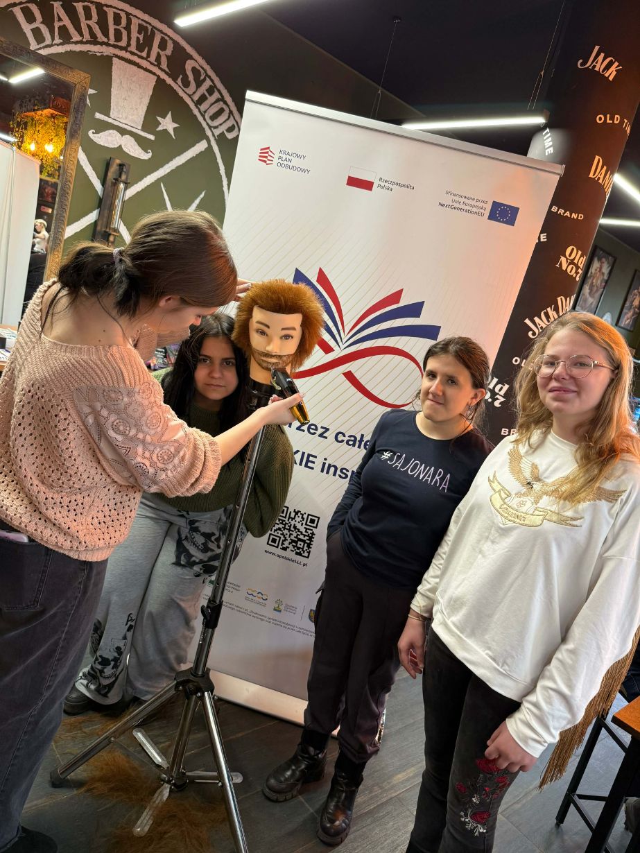 Grupa czterech młodych kobiet pozuje przy białym banerze z napisem „Nauka przez całe życie”. Jedna z nich trzyma maszynkę fryzjerską przy głowie treningowej zamontowanej na statywie, imitując strzyżenie. Na podłodze leżą ścięte kosmyki włosów, a w tle widoczne jest logo „Barber Shop”.