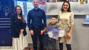 Michał Federer trzyma swoją pracę plastyczną z motywem księżyca oraz dyplom. Obok niego stoi kobieta w jasnym golfie i wzorzystej spódnicy, która trzyma go za ramię, a po lewej stronie zdjęcia widoczni są ci sami dwoje dorosłych (kobieta w żakiecie i mężczyzna w koszuli).