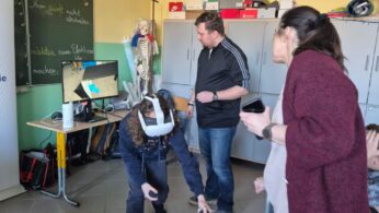 Instruktor oraz kobieta w bordowym kardiganie nadzorują ucznia, który schyla się podczas korzystania z gogli VR. Na ekranie monitora widoczna jest wirtualna szafka z niebieską strzałką wskazującą kierunek działania. Scena ma miejsce w dobrze oświetlonej sali lekcyjnej.
