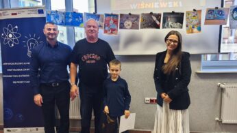 Mały chłopiec z torbą upominkową stoi w centrum między mężczyzną w niebieskiej koszuli, wysokim mężczyzną w czarnym t-shircie z napisem „CHICAGO” oraz kobietą w czarnym żakiecie i plisowanej spódnicy. Na ścianie za nimi wiszą prace plastyczne.