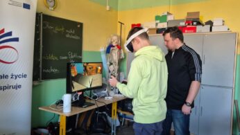 Uczeń w jasnozielonej bluzie testuje technologię VR pod okiem instruktora. Na monitorze widać podgląd wirtualnego biura lub pracowni z czarnym biurkiem. Po lewej stronie kadru widoczny jest fragment dużego białego baneru z logotypem unijnym.