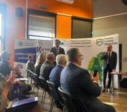 Dynamiczne ujęcie z sali konferencyjnej, na którym widać uczestników bijących brawo prelegentowi stojącemu przy stoliku. Na scenie znajduje się dwóch mężczyzn, z których jeden przemawia przez mikrofon. W tle widoczne są liczne banery organizacji takich jak Opolska Izba Gospodarcza czy PCK. Pomieszczenie charakteryzuje się intensywnie pomarańczową ścianą i nowoczesnym oświetleniem punktowym.