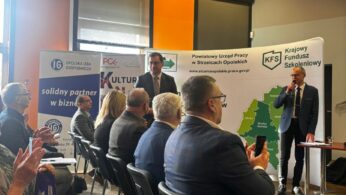 Dynamiczne ujęcie z sali konferencyjnej, na którym widać uczestników bijących brawo prelegentowi stojącemu przy stoliku. Na scenie znajduje się dwóch mężczyzn, z których jeden przemawia przez mikrofon. W tle widoczne są liczne banery organizacji takich jak Opolska Izba Gospodarcza czy PCK. Pomieszczenie charakteryzuje się intensywnie pomarańczową ścianą i nowoczesnym oświetleniem punktowym.