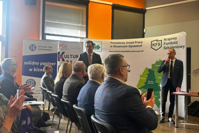 Dynamiczne ujęcie z sali konferencyjnej, na którym widać uczestników bijących brawo prelegentowi stojącemu przy stoliku. Na scenie znajduje się dwóch mężczyzn, z których jeden przemawia przez mikrofon. W tle widoczne są liczne banery organizacji takich jak Opolska Izba Gospodarcza czy PCK. Pomieszczenie charakteryzuje się intensywnie pomarańczową ścianą i nowoczesnym oświetleniem punktowym.