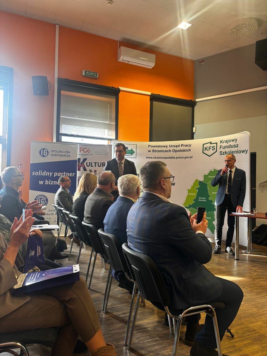 Dynamiczne ujęcie z sali konferencyjnej, na którym widać uczestników bijących brawo prelegentowi stojącemu przy stoliku. Na scenie znajduje się dwóch mężczyzn, z których jeden przemawia przez mikrofon. W tle widoczne są liczne banery organizacji takich jak Opolska Izba Gospodarcza czy PCK. Pomieszczenie charakteryzuje się intensywnie pomarańczową ścianą i nowoczesnym oświetleniem punktowym.