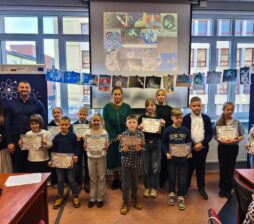 Grupowe zdjęcie przedstawia dzieci i dwoje dorosłych stojących w jasnej sali podczas uroczystości wręczenia dyplomów. Uczestnicy trzymają kolorowe certyfikaty, a w tle widać ekran z pracami plastycznymi o tematyce kosmicznej oraz flagi Unii Europejskiej i Polski. Dzieci są uśmiechnięte i ubrane w stroje galowe lub codzienne, a nad nimi rozwieszono sznur z ich rysunkami. Całość dokumentuje szkolne lub pozalekcyjne wydarzenie edukacyjne związane z konkursem artystycznym.