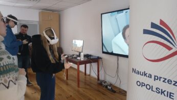 Na pierwszym planie widać kobietę w goglach VR, która za pomocą kontrolerów wchodzi w interakcję z wirtualnym otoczeniem. Po prawej stronie znajduje się baner z logotypem Krajowego Planu Odbudowy i hasłem „Opolskie inspiruje”. Na dużym ekranie w tle wyświetlany jest podgląd z gogli, przedstawiający wnętrze białego pomieszczenia.