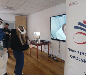 Na pierwszym planie widać kobietę w goglach VR, która za pomocą kontrolerów wchodzi w interakcję z wirtualnym otoczeniem. Po prawej stronie znajduje się baner z logotypem Krajowego Planu Odbudowy i hasłem „Opolskie inspiruje”. Na dużym ekranie w tle wyświetlany jest podgląd z gogli, przedstawiający wnętrze białego pomieszczenia.