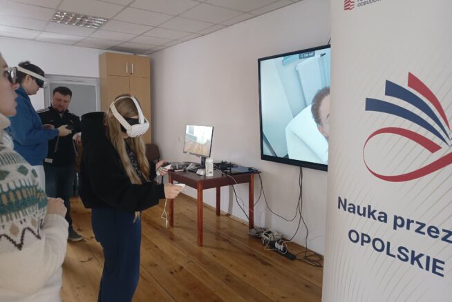 Na pierwszym planie widać kobietę w goglach VR, która za pomocą kontrolerów wchodzi w interakcję z wirtualnym otoczeniem. Po prawej stronie znajduje się baner z logotypem Krajowego Planu Odbudowy i hasłem „Opolskie inspiruje”. Na dużym ekranie w tle wyświetlany jest podgląd z gogli, przedstawiający wnętrze białego pomieszczenia.