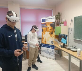 Dwóch uczniów na pierwszym planie korzysta z zestawów do rzeczywistości wirtualnej (gogle i kontrolery). W tle widoczne są stanowiska komputerowe, baner "Europe Direct" oraz szafki z roślinami doniczkowymi.