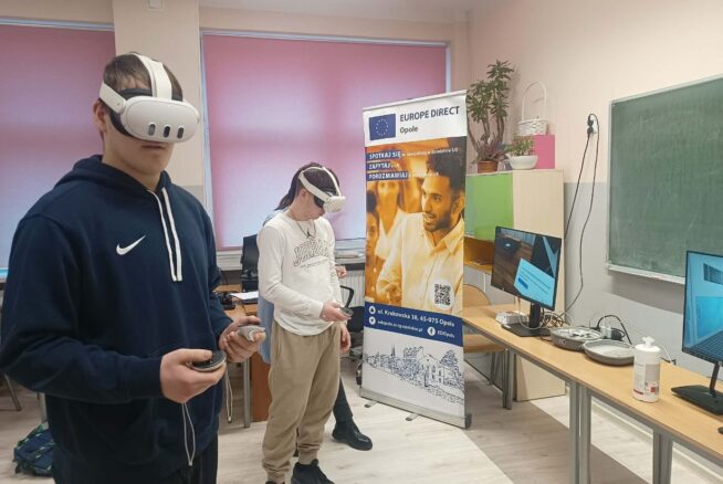 Dwóch uczniów na pierwszym planie korzysta z zestawów do rzeczywistości wirtualnej (gogle i kontrolery). W tle widoczne są stanowiska komputerowe, baner "Europe Direct" oraz szafki z roślinami doniczkowymi.