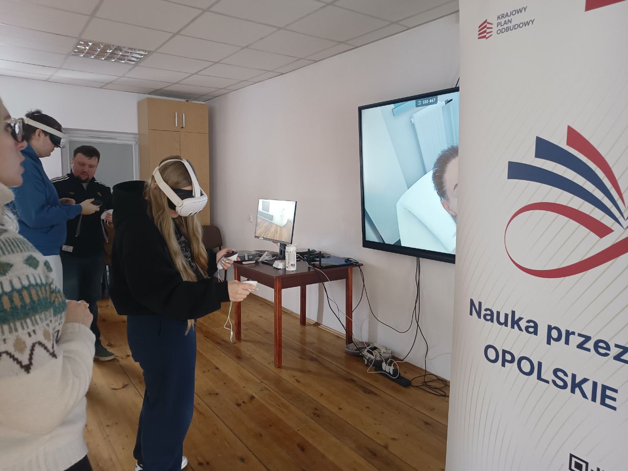 Na pierwszym planie widać kobietę w goglach VR, która za pomocą kontrolerów wchodzi w interakcję z wirtualnym otoczeniem. Po prawej stronie znajduje się baner z logotypem Krajowego Planu Odbudowy i hasłem „Opolskie inspiruje”. Na dużym ekranie w tle wyświetlany jest podgląd z gogli, przedstawiający wnętrze białego pomieszczenia.