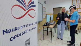 Kobieta w okularach i czarnej marynarce pomaga młodemu chłopakowi w goglach VR w obsłudze bezprzewodowych kontrolerów. W tle widoczna jest sala lekcyjna z tablicami informacyjnymi oraz biurko ze sprzętem komputerowym. Na pierwszym planie po lewej stronie dominuje duży biały baner z kolorową grafiką i kodem QR.