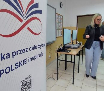 Kobieta w okularach i czarnej marynarce pomaga młodemu chłopakowi w goglach VR w obsłudze bezprzewodowych kontrolerów. W tle widoczna jest sala lekcyjna z tablicami informacyjnymi oraz biurko ze sprzętem komputerowym. Na pierwszym planie po lewej stronie dominuje duży biały baner z kolorową grafiką i kodem QR.
