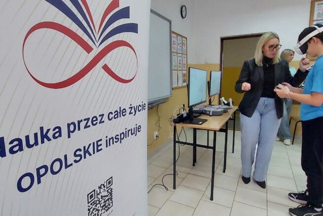 Kobieta w okularach i czarnej marynarce pomaga młodemu chłopakowi w goglach VR w obsłudze bezprzewodowych kontrolerów. W tle widoczna jest sala lekcyjna z tablicami informacyjnymi oraz biurko ze sprzętem komputerowym. Na pierwszym planie po lewej stronie dominuje duży biały baner z kolorową grafiką i kodem QR.