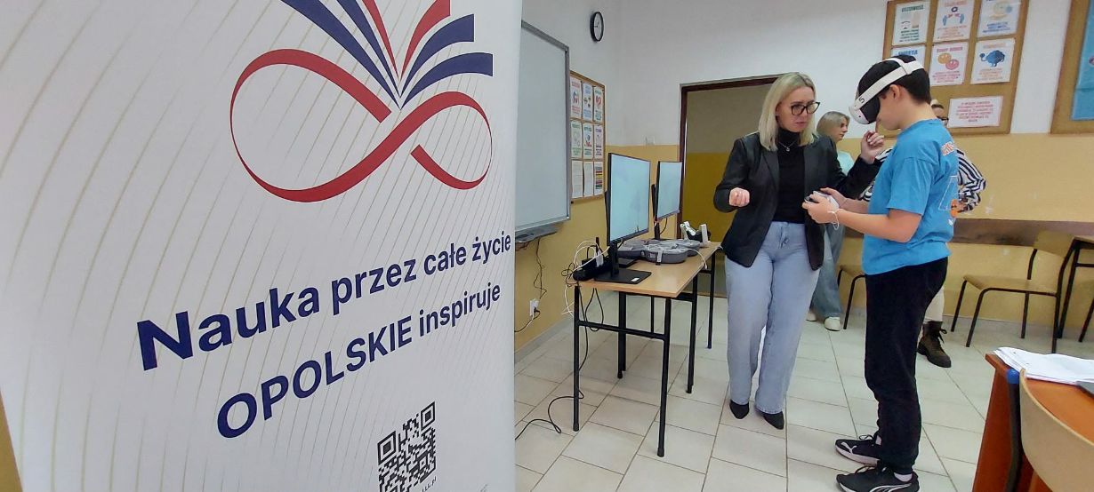 Kobieta w okularach i czarnej marynarce pomaga młodemu chłopakowi w goglach VR w obsłudze bezprzewodowych kontrolerów. W tle widoczna jest sala lekcyjna z tablicami informacyjnymi oraz biurko ze sprzętem komputerowym. Na pierwszym planie po lewej stronie dominuje duży biały baner z kolorową grafiką i kodem QR.