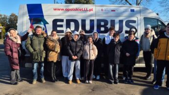 Szerokie ujęcie grupy uczniów i opiekunów stojących przed furgonem „EDUBUS”, który służy jako mobilne centrum edukacyjne. Wszyscy są ubrani w odzież wierzchnią, a na boku pojazdu widnieje postać robota w birecie oraz hasła promujące edukację w województwie opolskim.