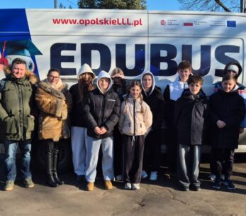 Szerokie ujęcie grupy uczniów i opiekunów stojących przed furgonem „EDUBUS”, który służy jako mobilne centrum edukacyjne. Wszyscy są ubrani w odzież wierzchnią, a na boku pojazdu widnieje postać robota w birecie oraz hasła promujące edukację w województwie opolskim.