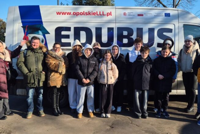 Szerokie ujęcie grupy uczniów i opiekunów stojących przed furgonem „EDUBUS”, który służy jako mobilne centrum edukacyjne. Wszyscy są ubrani w odzież wierzchnią, a na boku pojazdu widnieje postać robota w birecie oraz hasła promujące edukację w województwie opolskim.