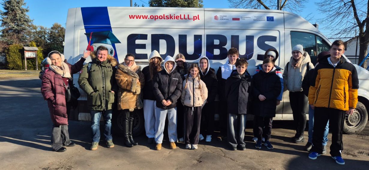 Szerokie ujęcie grupy uczniów i opiekunów stojących przed furgonem „EDUBUS”, który służy jako mobilne centrum edukacyjne. Wszyscy są ubrani w odzież wierzchnią, a na boku pojazdu widnieje postać robota w birecie oraz hasła promujące edukację w województwie opolskim.