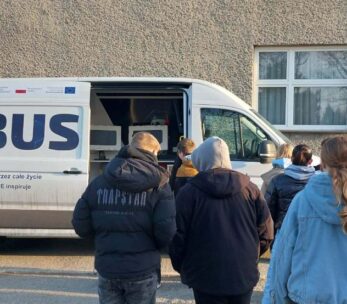 Grupa młodych ludzi stoi przed białym busem edukacyjnym z napisem „Nauka przez całe życie – OPOLSKIE inspiruje”. Drzwi przesuwne pojazdu są otwarte, odsłaniając wnętrze wyposażone w sprzęt elektroniczny. Akcja dzieje się na zewnątrz, przy szarym budynku w słoneczny dzień.