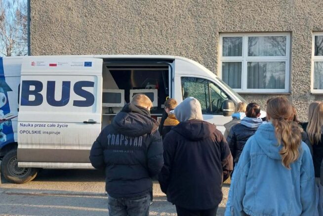 Grupa młodych ludzi stoi przed białym busem edukacyjnym z napisem „Nauka przez całe życie – OPOLSKIE inspiruje”. Drzwi przesuwne pojazdu są otwarte, odsłaniając wnętrze wyposażone w sprzęt elektroniczny. Akcja dzieje się na zewnątrz, przy szarym budynku w słoneczny dzień.