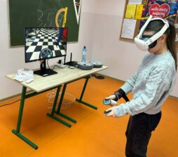 Uczennica w szarym, plecionym swetrze stoi z profilu, mając założone białe gogle VR i trzymając kontrolery w obu dłoniach. Przed nią na stoliku stoi monitor wyświetlający wnętrze warsztatu samochodowego (widoczna opona i szachownica na podłodze). W tle znajduje się zielona tablica z przyborami geometrycznymi oraz plakat WOŚP.