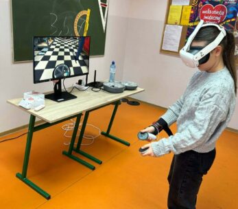 Uczennica w szarym, plecionym swetrze stoi z profilu, mając założone białe gogle VR i trzymając kontrolery w obu dłoniach. Przed nią na stoliku stoi monitor wyświetlający wnętrze warsztatu samochodowego (widoczna opona i szachownica na podłodze). W tle znajduje się zielona tablica z przyborami geometrycznymi oraz plakat WOŚP.