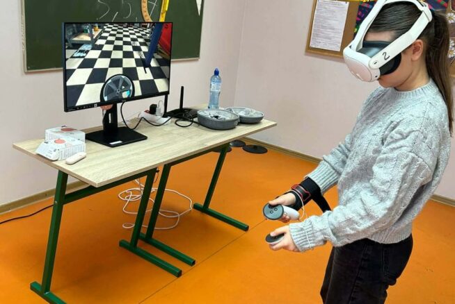 Uczennica w szarym, plecionym swetrze stoi z profilu, mając założone białe gogle VR i trzymając kontrolery w obu dłoniach. Przed nią na stoliku stoi monitor wyświetlający wnętrze warsztatu samochodowego (widoczna opona i szachownica na podłodze). W tle znajduje się zielona tablica z przyborami geometrycznymi oraz plakat WOŚP.