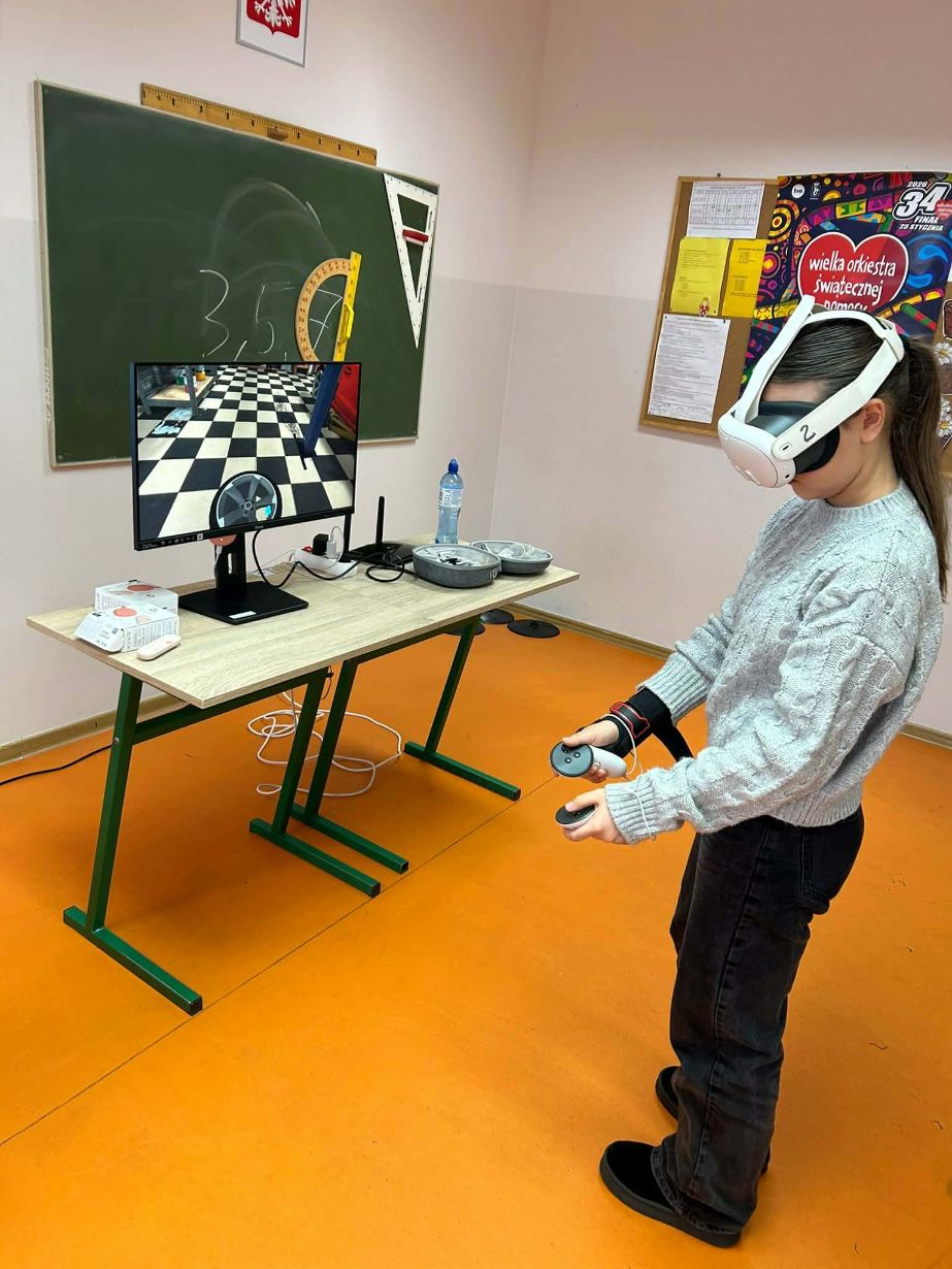Uczennica w szarym, plecionym swetrze stoi z profilu, mając założone białe gogle VR i trzymając kontrolery w obu dłoniach. Przed nią na stoliku stoi monitor wyświetlający wnętrze warsztatu samochodowego (widoczna opona i szachownica na podłodze). W tle znajduje się zielona tablica z przyborami geometrycznymi oraz plakat WOŚP.