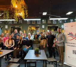 Grupa około dwudziestu osób, w tym młodzież i dorośli, pozuje do wspólnego zdjęcia wewnątrz stylowego salonu fryzjerskiego. Po prawej stronie stoi mężczyzna w szarej marynarce obok dużego baneru promującego projekt edukacyjny w województwie opolskim. Wnętrze udekorowane jest wiszącymi roślinami, ozdobnymi żarówkami i rzędami grafik na ścianach.