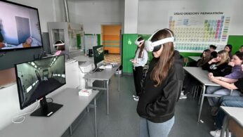 Szerokie ujęcie sali lekcyjnej, w której grupa uczniów testuje technologię VR pod nadzorem rówieśników i nauczycieli. Na pierwszym planie dziewczyna w ciemnej bluzie i jeansach korzysta z gogli, a na monitorach obok widać symulacje wnętrz budynków. Ściany klasy zdobi duży układ okresowy pierwiastków oraz pomoce naukowe, co podkreśla naukowy charakter zajęć. Reszta klasy siedzi w ławkach, obserwując przebieg ćwiczeń wirtualnych.