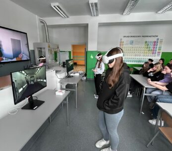 Szerokie ujęcie sali lekcyjnej, w której grupa uczniów testuje technologię VR pod nadzorem rówieśników i nauczycieli. Na pierwszym planie dziewczyna w ciemnej bluzie i jeansach korzysta z gogli, a na monitorach obok widać symulacje wnętrz budynków. Ściany klasy zdobi duży układ okresowy pierwiastków oraz pomoce naukowe, co podkreśla naukowy charakter zajęć. Reszta klasy siedzi w ławkach, obserwując przebieg ćwiczeń wirtualnych.