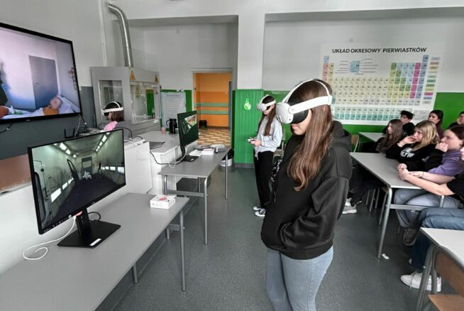 Szerokie ujęcie sali lekcyjnej, w której grupa uczniów testuje technologię VR pod nadzorem rówieśników i nauczycieli. Na pierwszym planie dziewczyna w ciemnej bluzie i jeansach korzysta z gogli, a na monitorach obok widać symulacje wnętrz budynków. Ściany klasy zdobi duży układ okresowy pierwiastków oraz pomoce naukowe, co podkreśla naukowy charakter zajęć. Reszta klasy siedzi w ławkach, obserwując przebieg ćwiczeń wirtualnych.