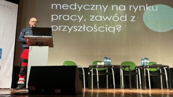 Mężczyzna w mundurze ratownika medycznego przemawia zza mównicy na scenie. W tle widać slajd z napisem „Ratownik medyczny na rynku pracy, zawód z przyszłością?”. Obok mównicy stoją puste zielone krzesła i stoliki z wodą mineralną.