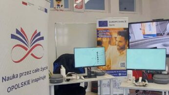 Wnętrze nowoczesnej sali edukacyjnej w Opolu. Na pierwszym planie po lewej stronie widoczny jest pionowy baner z logotypem splecionych kolorowych linii i napisem „Nauka przez całe życie OPOLSKIE inspiruje”. W głębi znajdują się dwa biurka z monitorami komputerowymi, goglami VR oraz kontrolerami. W tle widoczny jest baner punktu Europe Direct Opole oraz okna z dziecięcymi rysunkami i godłem Polski.