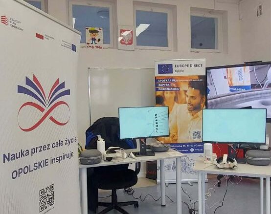 Wnętrze nowoczesnej sali edukacyjnej w Opolu. Na pierwszym planie po lewej stronie widoczny jest pionowy baner z logotypem splecionych kolorowych linii i napisem „Nauka przez całe życie OPOLSKIE inspiruje”. W głębi znajdują się dwa biurka z monitorami komputerowymi, goglami VR oraz kontrolerami. W tle widoczny jest baner punktu Europe Direct Opole oraz okna z dziecięcymi rysunkami i godłem Polski.