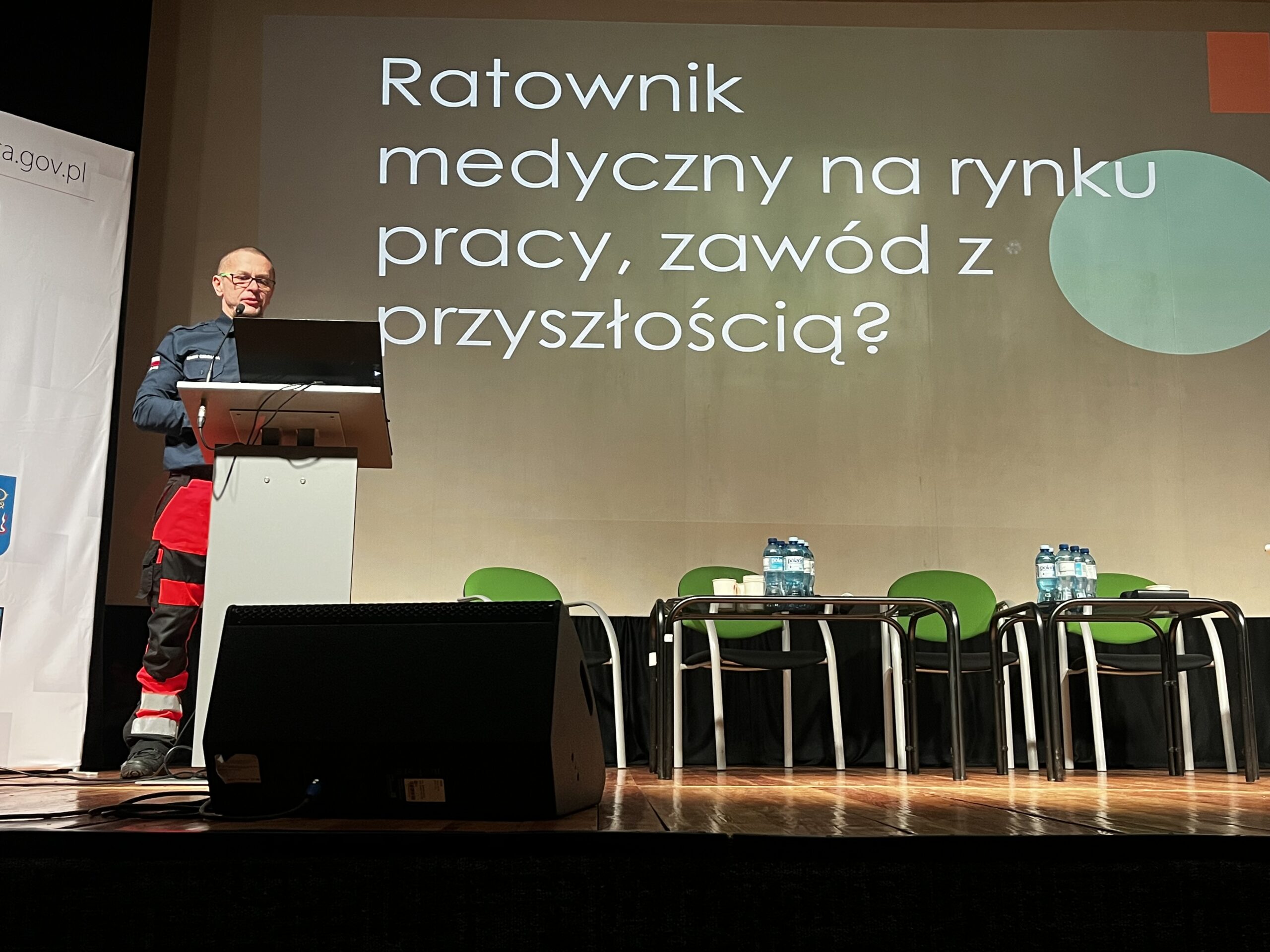 Mężczyzna w mundurze ratownika medycznego przemawia zza mównicy na scenie. W tle widać slajd z napisem „Ratownik medyczny na rynku pracy, zawód z przyszłością?”. Obok mównicy stoją puste zielone krzesła i stoliki z wodą mineralną.