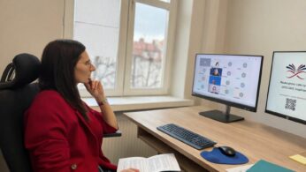 Kobieta w czerwonej marynarce siedzi przy biurku i z uwagą obserwuje dwa monitory, na których widać wideokonferencję oraz prezentację. W dłoniach trzyma notatki, a w tle znajduje się okno z widokiem na dachy budynków. Całość przedstawia profesjonalne stanowisko pracy podczas zdalnego spotkania.