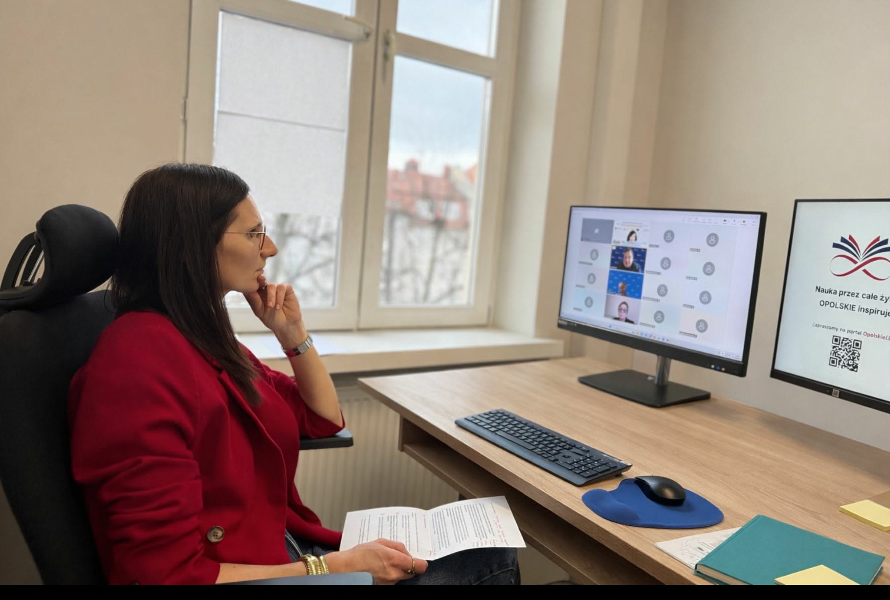 Kobieta w czerwonej marynarce siedzi przy biurku i z uwagą obserwuje dwa monitory, na których widać wideokonferencję oraz prezentację. W dłoniach trzyma notatki, a w tle znajduje się okno z widokiem na dachy budynków. Całość przedstawia profesjonalne stanowisko pracy podczas zdalnego spotkania.