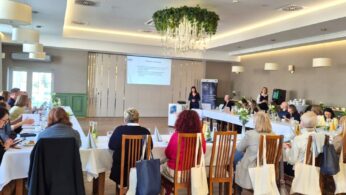 Szerokie ujęcie sali konferencyjnej, w której grupa osób siedzi przy stołach ustawionych w podkowę, słuchając prelegentki stojącej przy ekranie. Wnętrze jest jasne, z eleganckim żyrandolem ozdobionym zielenią i zastawionym bufetem w tle. Na ekranie widoczny jest slajd z napisem „Praktyki zawodowe”.