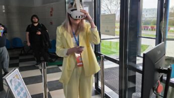 Kobieta w żółtym garniturze stoi przy stanowisku informacyjnym i testuje białe gogle VR, trzymając w dłoni kontroler. W tle widać przeszklone wejście do budynku oraz biurko z laptopem i monitorem. Podłoga w pomieszczeniu wyłożona jest czarno-białymi płytkami w szachownicę.